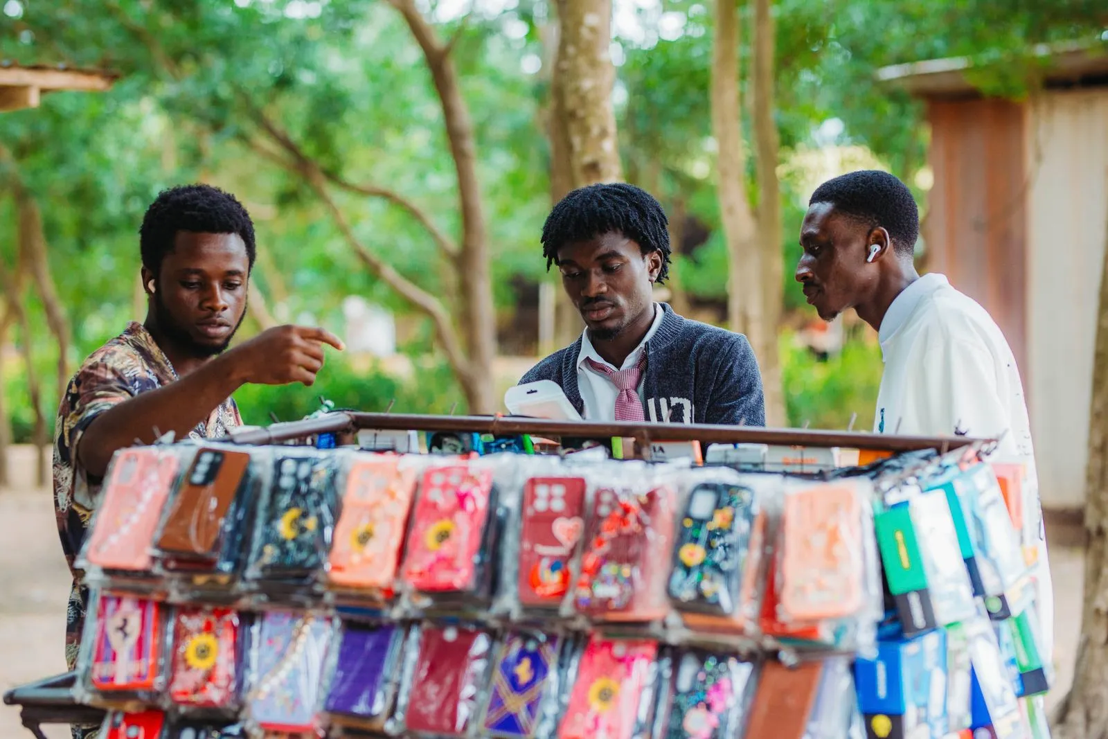 Two customers at an outdoor market stall looking at a phone with the trader, choosing from his rack of phone cases
